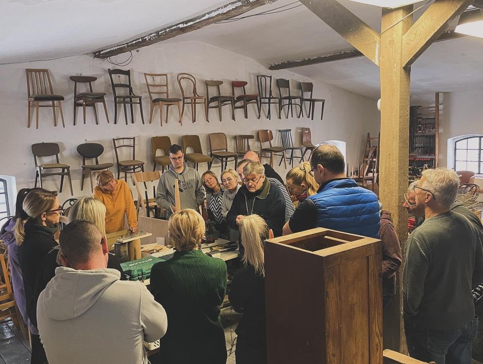 Obraz przedstawia grupę ludzi uważnie dyskutujących lub badających jakiś obiekt w warsztacie lub muzeum. Pokój wydaje się być warsztatem lub wycieczką. Ściany ozdobione są rzędami krzeseł o różnych wzorach i kolorach, podkreślając temat miejsca, być może związany z projektowaniem mebli lub rzemiosłem. Przestrzeń ma nieco antyczny wygląd z widocznymi drewnianymi belkami i tynkiem na ścianach. Oświetlenie jest miękkie, tworząc przytulną atmosferę.