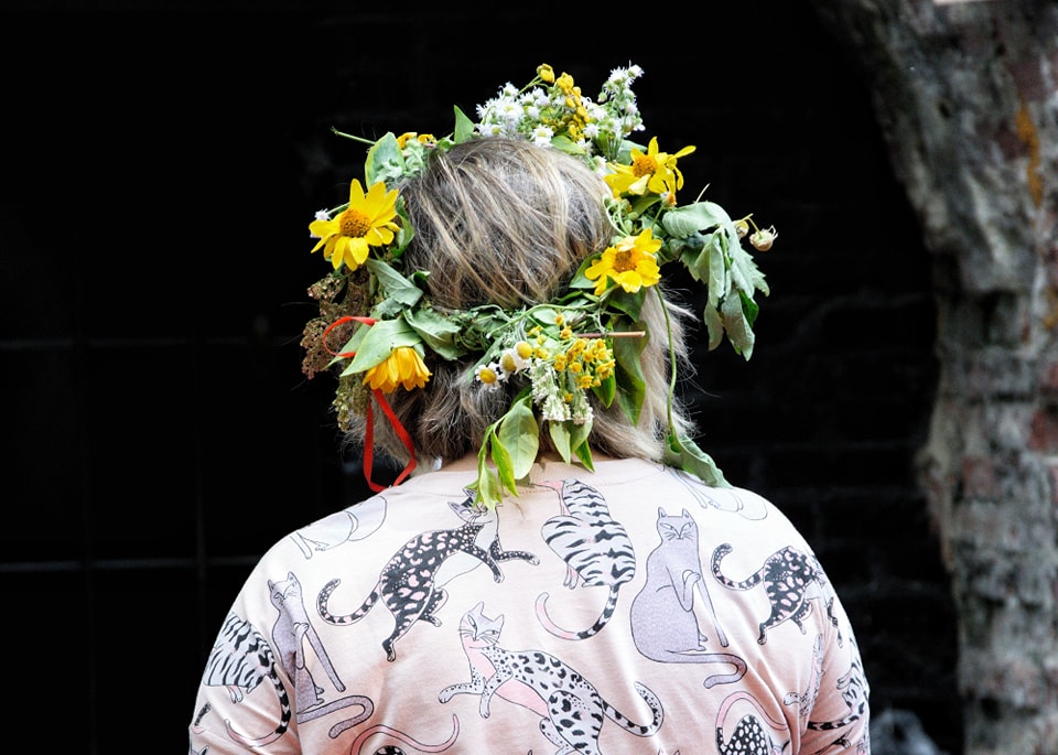 Osoba odwrócona tyłem. Na głowie ma wianek z żółtych i polnych kwiatów. Na sobie ma różową koszulę z printem w koty.