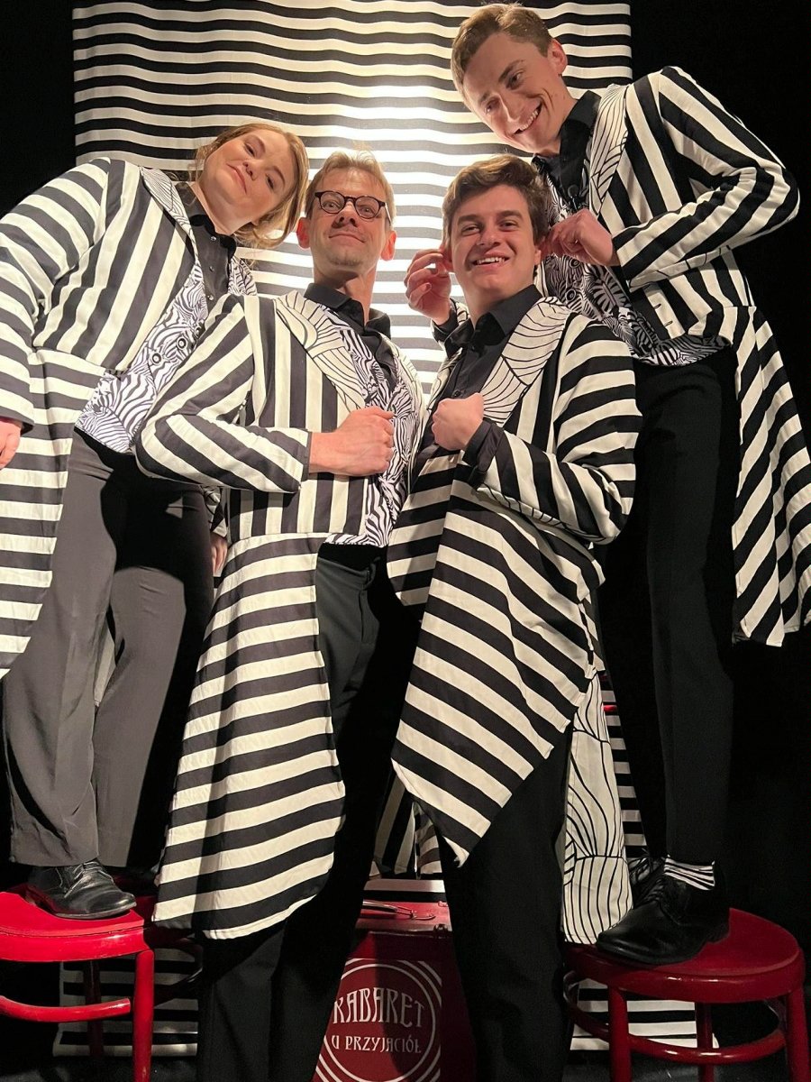 Zdjęcie aktorów - kobieta i trzech mężczyzn we frakach w biało-czarne paski. Stoją obok siebie, wpatrzeni w obiektyw, uśmiechają się.