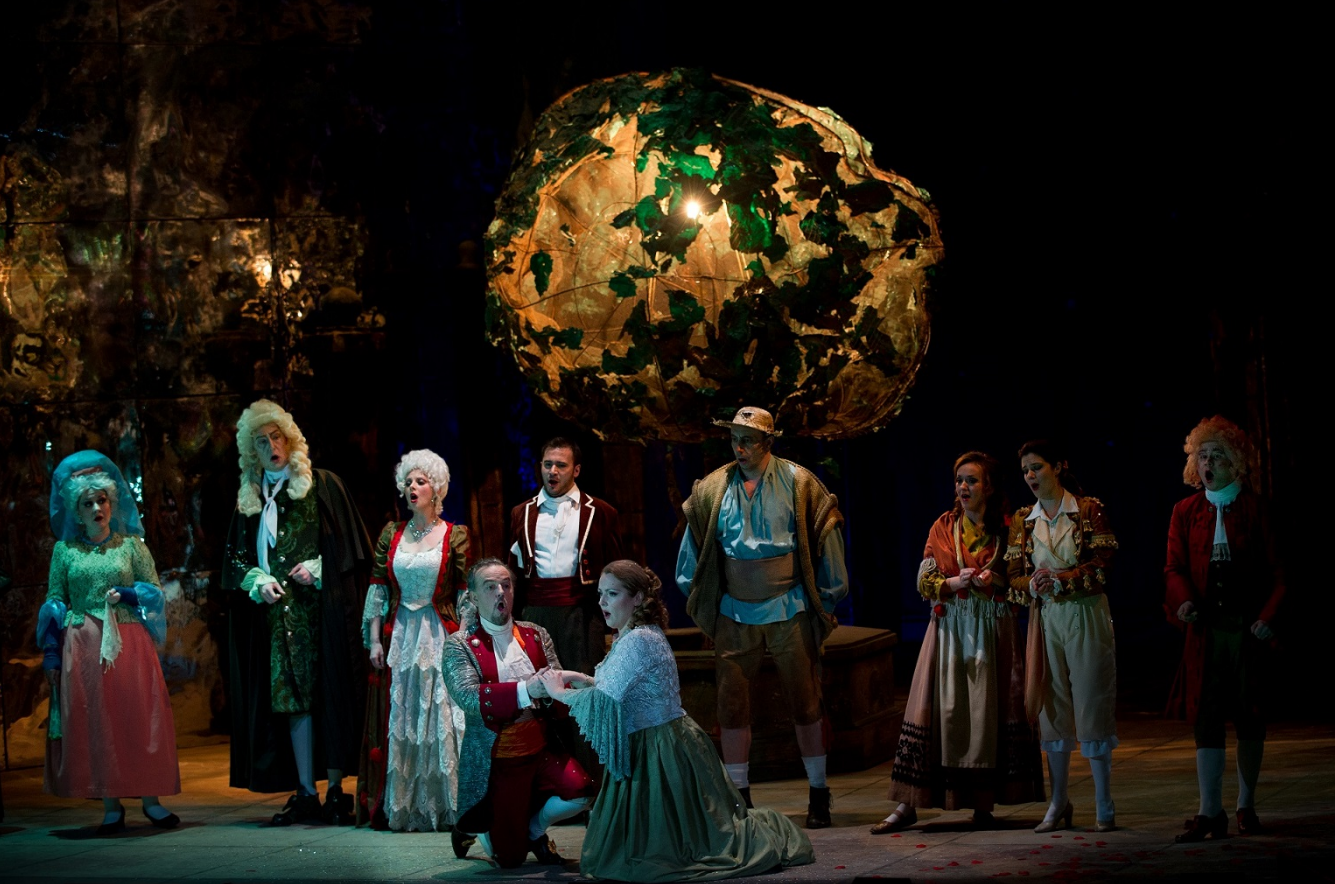 Na scenie aktorzy w kostiumach z epoki, na pierwszym planie klęczą mężczyzna i kobieta, za nimi rząd innych postaci. Wszyscy śpiewają.