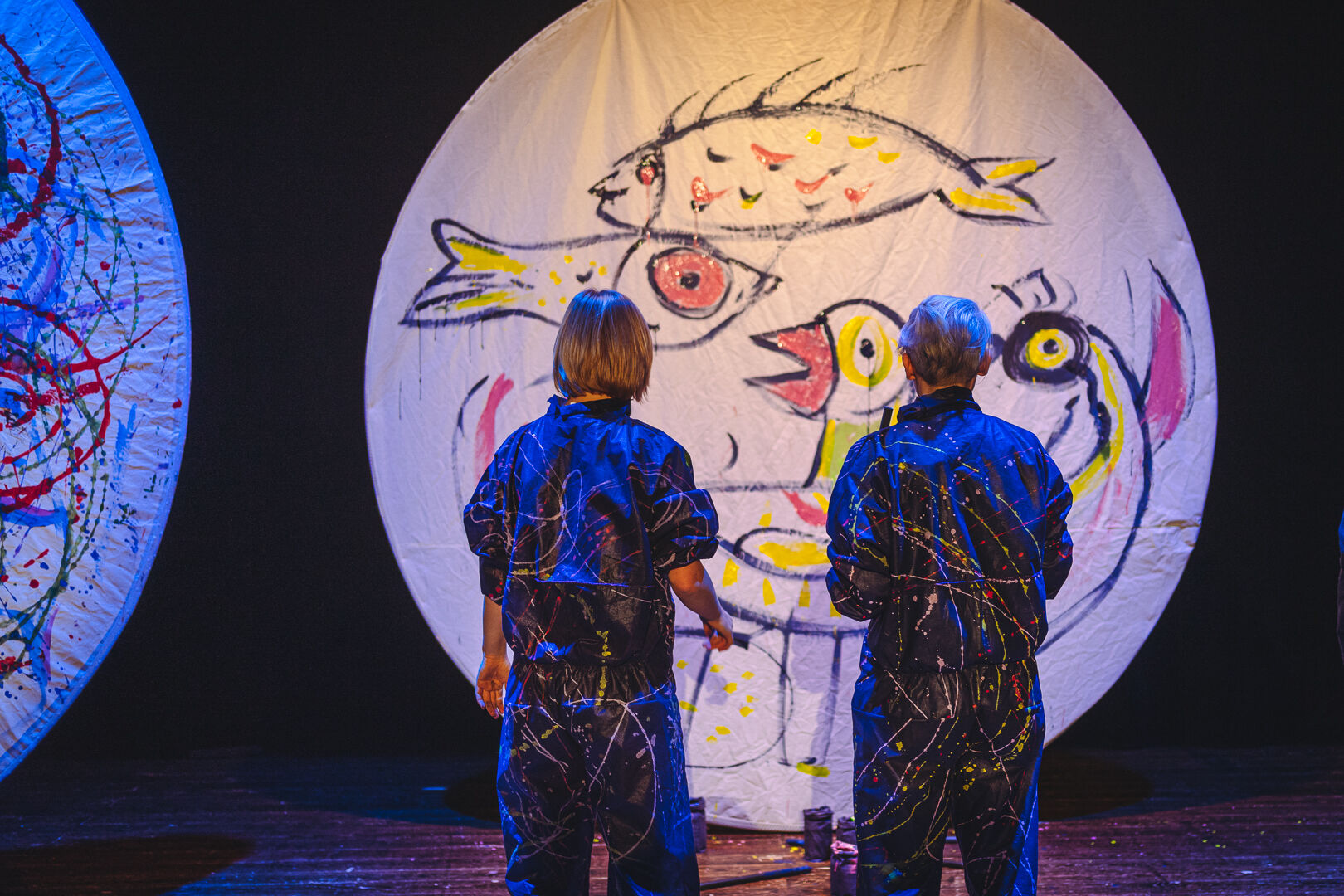 na zdjeciu widoczne dwie osoby stojące tyłem, w granatowych kombinezonach popisanych kolorowymi farbami, przed nimi biala płaszczyzna w ksztalcie koła, na której narysowano kolorowe ryby i ptaki