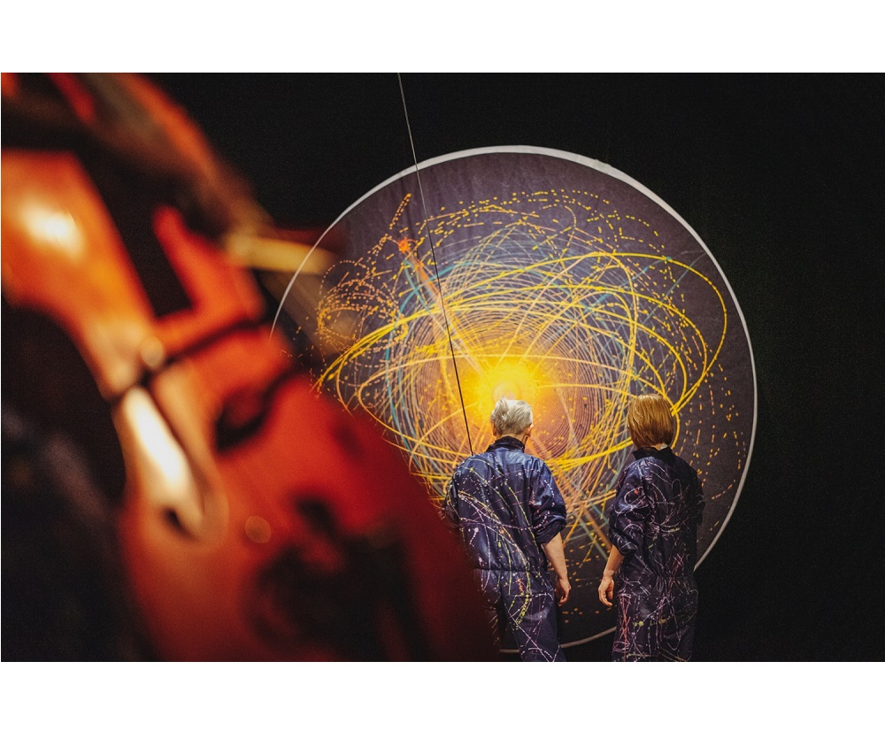 na zdjeciu widoczne dwie osoby stojące tyłem, w granatowych kombinezonach popisanych kolorowymi farbami, przed nimi czarna płaszczyzna w kształcie koła, na której narysowano kolorowe okręgi i elipsy