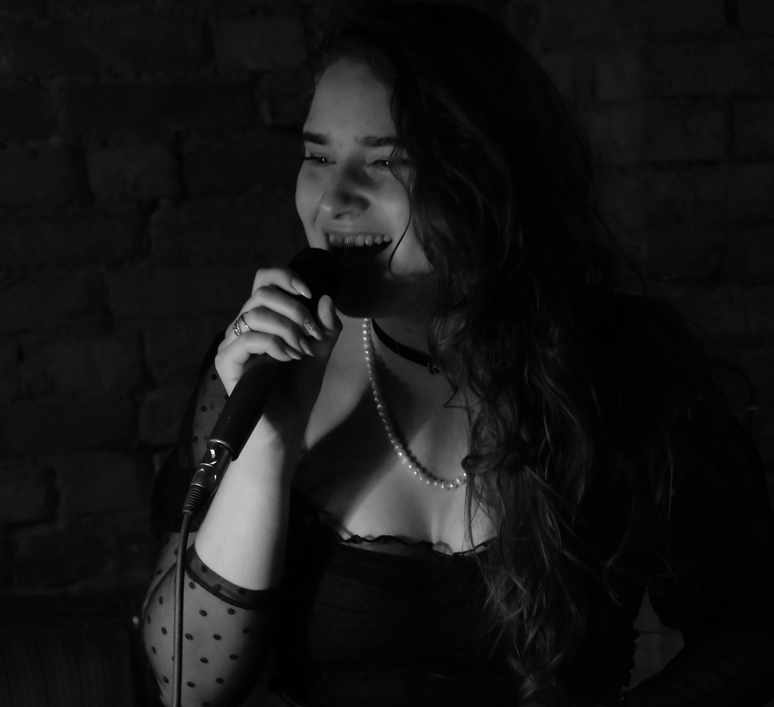 Zdjęcie, dziewczyna z mikrofonem w ciemnym pomieszczeniu. Dziewczyna ma ropuszczone długie włosy, koronkową bluzkę i naszyjnik z pereł.