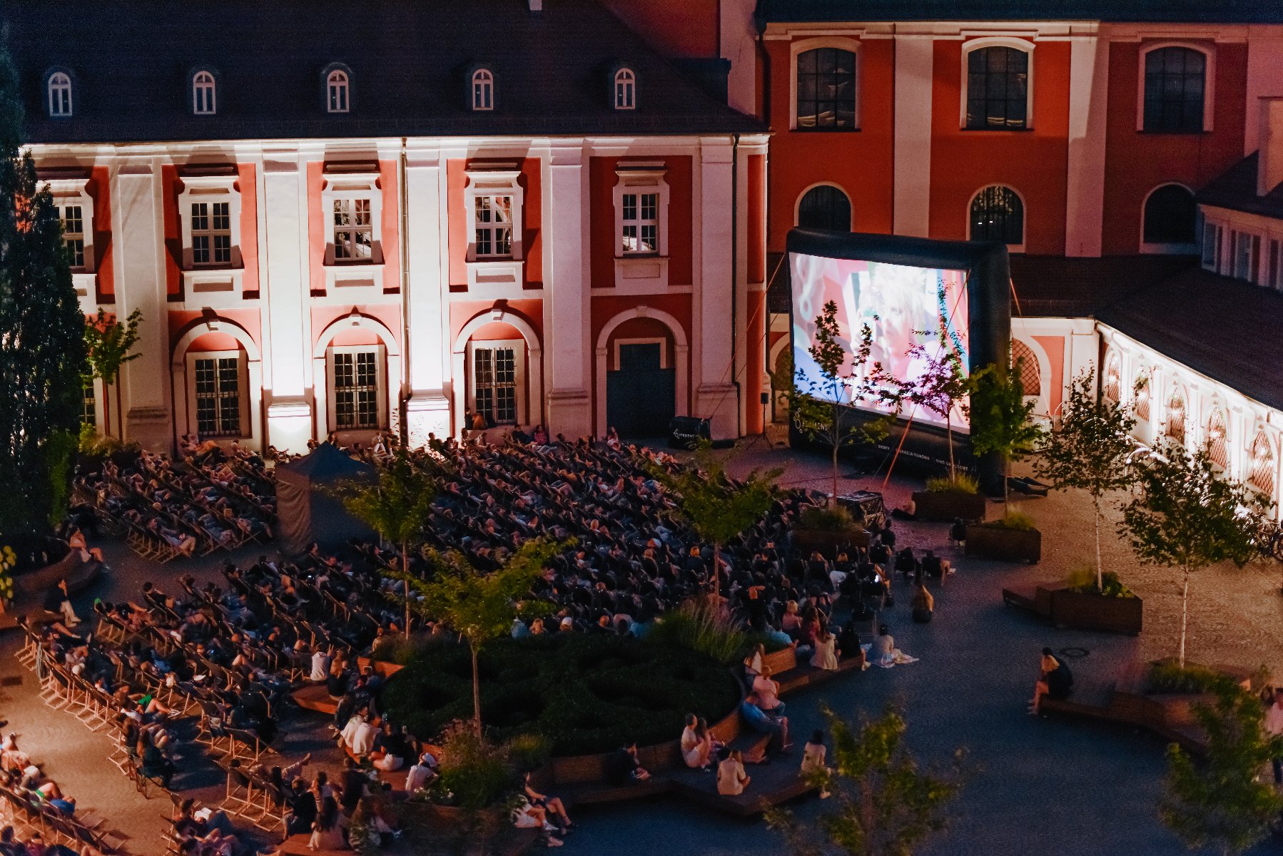 Pokaz filmowy na Wolnym Dziedzińcu. Na dziedzińcu Urzędu Miasta ogormny ekran kinowy, przed którym siedzą ludzie na leżakach i oglądają pokaz filmowy.