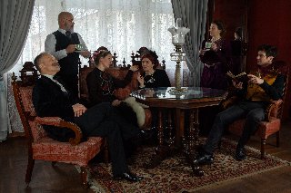 Zdjęcie, kadr filmu, aktorzy siedzą na eleganckich fotelach w salonie, w dłoniach trzymają filiżanki, przed nimi drewniany rzeźbiony stół.