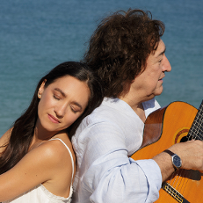 Kobieta i mężczyzna siedzą na plaży, odwróceni do siebie plecami. Mężczyzna gra na gitarze.