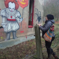 Kobieta patrzy na grafitti, namalowane na ścianie pustostanu. Grafitti przedstawia klauna Pennywise z filmu "To" na podstawie powieści Stephena Kinga. Klaun ma rozmiary dziecka, ale złowieszczy uśmiech, w ręku trzyma balonik, który przypomina dynię na Halloween - z wyciętymi oczami i ustami.