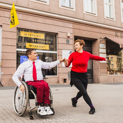 Kobieta w czerwonym sweterku i mężczyzna w białej koszuli i z czerwonym krawatem tańczą na ulicy. Mężczyzna porusza się na wózku inwalidzkim.