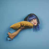 Kolażowe zdjęcie dziewczyny w niebieskich włosach przykładającej głowę do trzymanej przez rękę kogoś innego żółtej poduszki.