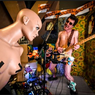"Bajzel". Nagi gitarzysta przy mikrofonie. Gitara zakrywa jego brzuch, częśći intymne i uda. Z przodu nagi manekin kobiety z zaklejonymi taśmą piersiami.