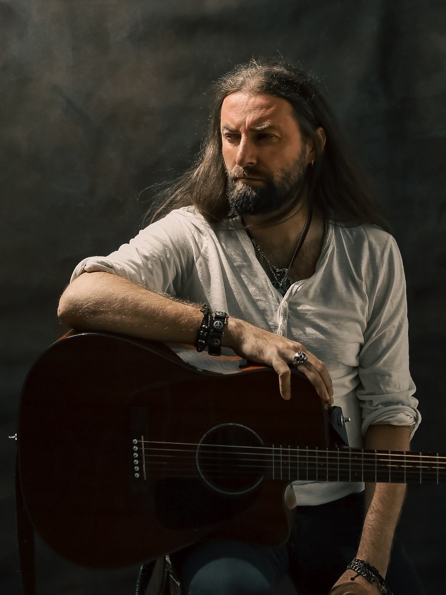 Mężczyzna w średnim wieku z długimi ciemnymi włosami i brodą. Ubrany w białą luźną koszulkę. Siedzi na wysokim krześle i jedną rękę opiera na gitarze akustycznej. Na ręce i na szyi ma biżuterie. Spogląda w bok.