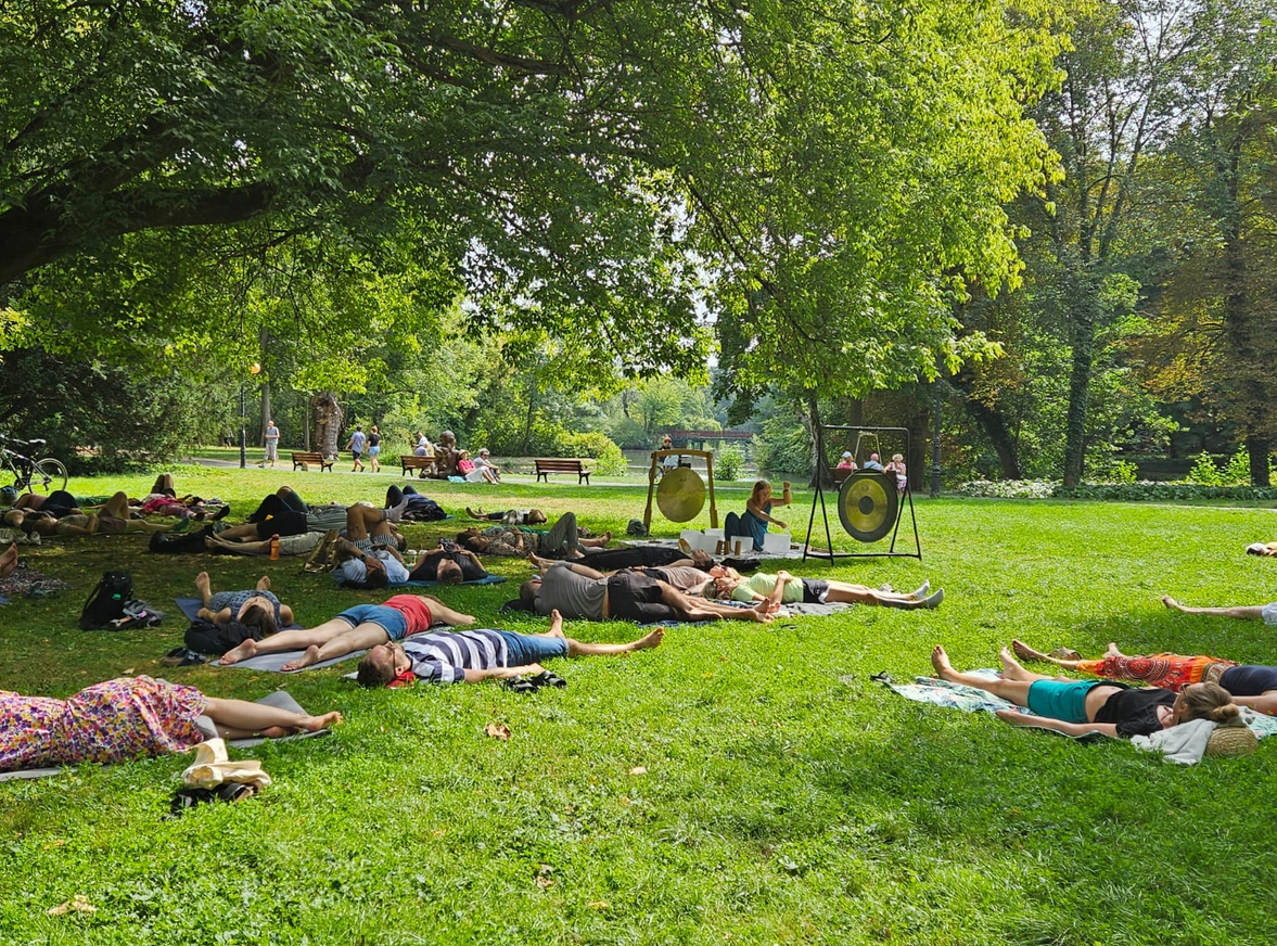 Zdjęcie, polana w parku, wokół drzewa, ludzie leżą na karimatach, przed nimi artyska gra na gongach i misach.