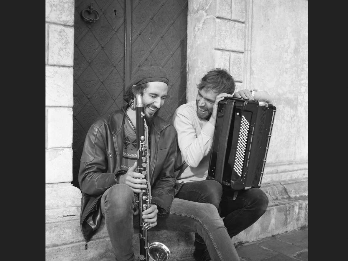Dwóch śmiejących się mężczyzn siedzi u drzwi kamienicy, jeden trzyma akordeon, drugi saksofon.