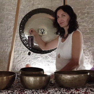Zdjęcie przedstawia kobietę siedzącą przed dużym gongiem oraz kilkoma misami do śpiewu. Kobieta trzyma mały, czarny, prostokątny przedmiot w swojej prawej ręce. Ma na sobie białą koszulkę bez rękawów i naszyjnik. Gong jest zawieszony na suficie za kobietą. Misy do śpiewu są ułożone w półkolu przed nią. Tło stanowi biała ściana. Kobieta ma ciemne włosy i patrzy w obiektyw aparatu. Zdjęcie wydaje się być wykonane w pomieszczeniu z białą ceglaną ścianą. Kobieta lekko się uśmiecha.