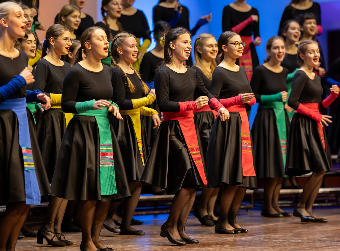 Chór dziewczęcy w czarnych sukienkach przepasanych kolorowymi wstęgami.