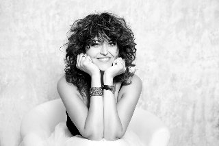 Czarno-białe zdjęcie przedstawia siedzącą i uśmiechającą się kobietę, która opiera swoją głowę na rękach wspartych łokciami o kolana.