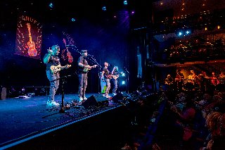 Zdjęcie przedstawia czterech muzyków występujących na scenie, grających na ukulele.