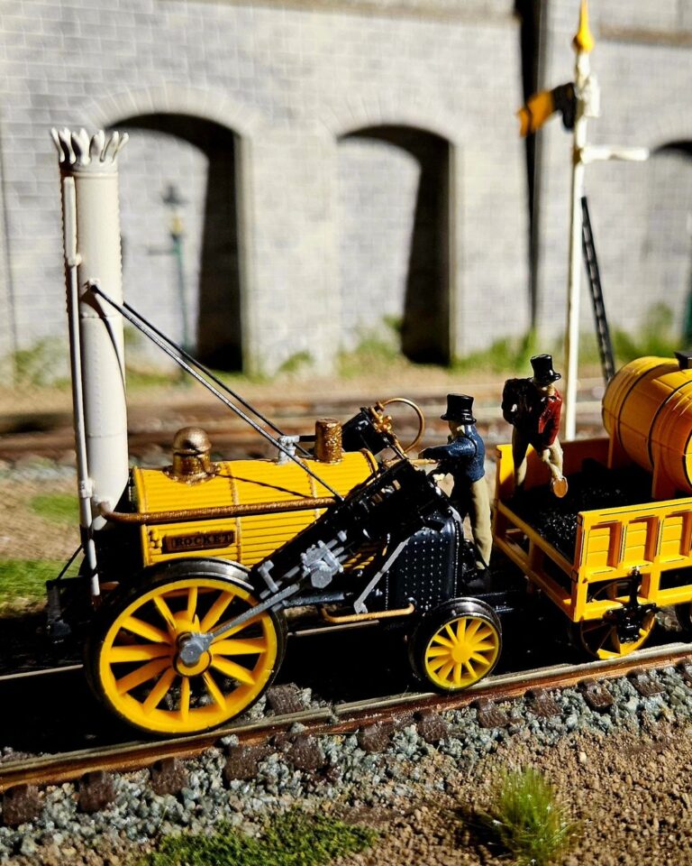 To jest miniaturowy model parowozu Rocket, historycznego pociągu, który zrewolucjonizował transport. Został ustawiony na makiecie torów kolejowych, kompletny z miniaturowym wagonem węglowym i figurkami ludzi. Model jest bardzo szczegółowy, z dokładnymi reprezentacjami projektu lokomotywy, w tym kotła, kół i nawet kominu. Model jest również pomalowany w jaskrawych kolorach, co dodaje realizmu. Tło zdjęcia wydaje się być miniaturowym modelem kamiennego muru, z arkadami w tle, dodającym do poczucia skali.