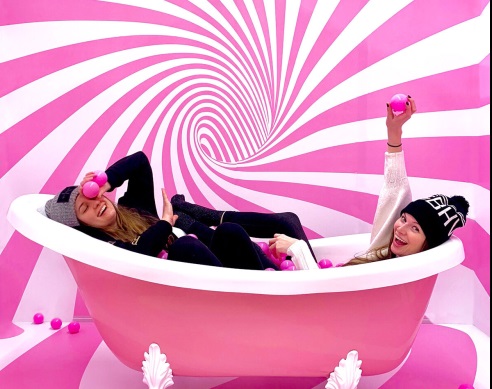 Dwie dziewczyny w ubraniu i w czapkach w różowej wannie. Na ścianie biało-różowy wir. W dłoniach maja różowe kulki. Dziewczyna po lewej ma wyprostowana do góry rękę.