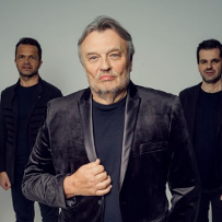 Krzysztof Cugowski jest ubrany w czarny garnitur z aksamitu, czarną koszulkę oraz spodnie. Za nim stoi czterech członków zespołu - wszyscy w czerni.