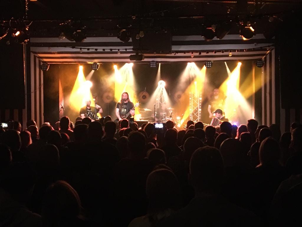 Na pierwszym planie głowy widowni, która słucha koncertu. Na oświetlonej scenie piątka mężczyzn w średnim wieku gra koncert na różnych instrumentach. Wśród nich jest gitarzysta, bębniarz, wiolonczelista, akordeonista.