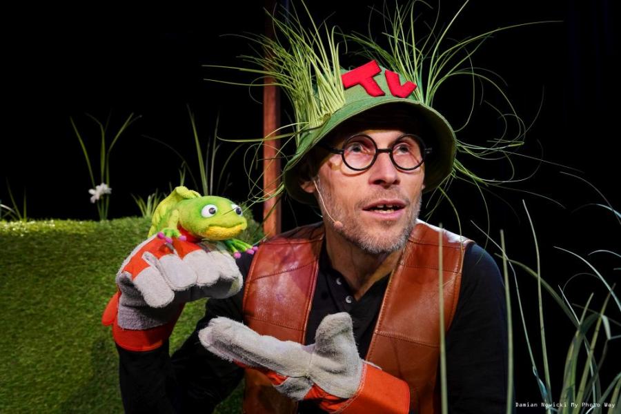 Zdjęcie aktora w zielonym kapeluszu, porośniętym trawami, który trzyma w ręce pacynkę żabę.