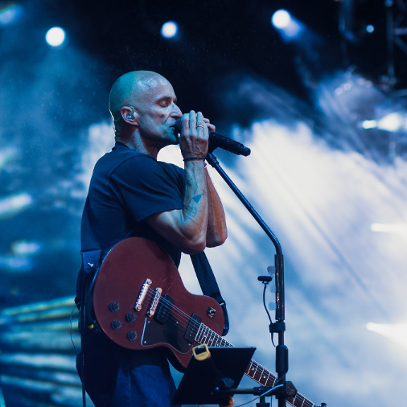 Łysy mężczyna przy mikrofonie, z gitarą na scenie.