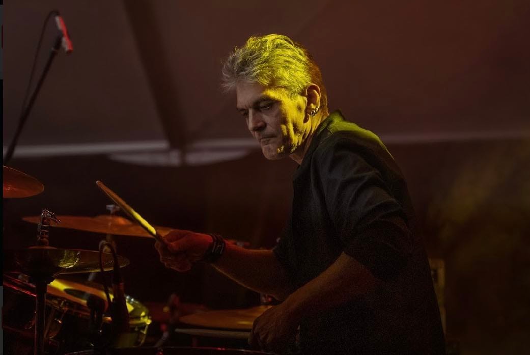 Mężczyzna w średnim wieku, z siwymi włosami i kolczykami w uchu. Ubrany w czarną koszulę z podwiniętymi rękawami. Mężczyzna gra na perkusji.