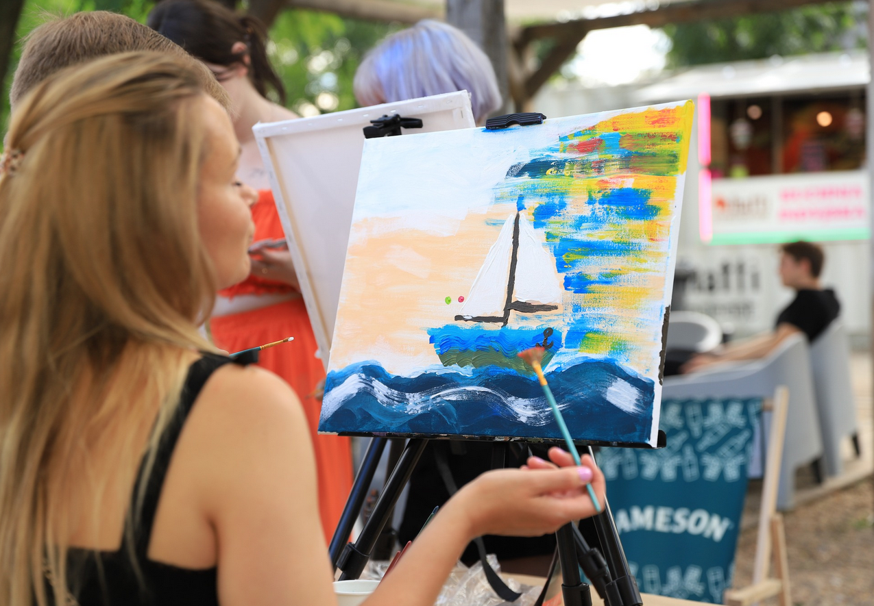 Zdjęcie, kobieta stoi przed malowanym przez siebie obrazem, na obrazie żagłówka.