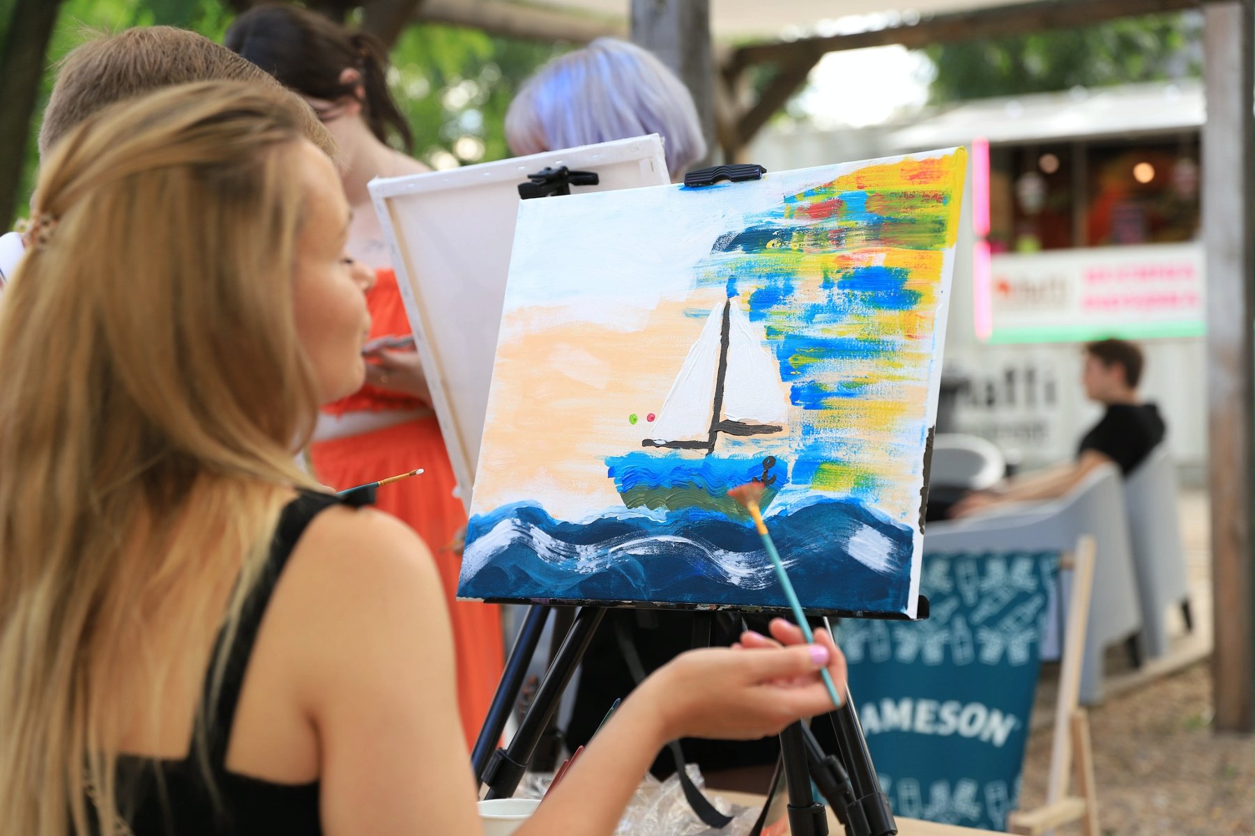 Kobieta zwrócona przodem do płótna. Maluje na nim obraz. który przedstawia łódkę na wodzie.