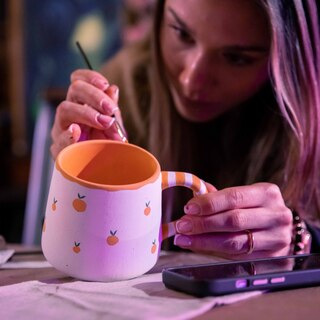 Kobieta w skupieniu maluje ceramiczny kubek cienkim pędzlem. Kubek jest biały, ozdobiony wzorem w małe pomarańcze, ma pomarańczowe wnętrze oraz ucho w biało-pomarańczowe paski. Kobieta trzyma naczynie lewą ręką, a obok niej na stole leży smartfon.