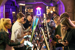 Grupa osób maluje obrazy, siedząc w pomieszczeniu o ceglanych ścianach.