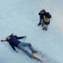 Martwe ciało leży na śniegu we krwi. Stoją nad nim dwie osoby, jedna z nich wykonuje telefon.