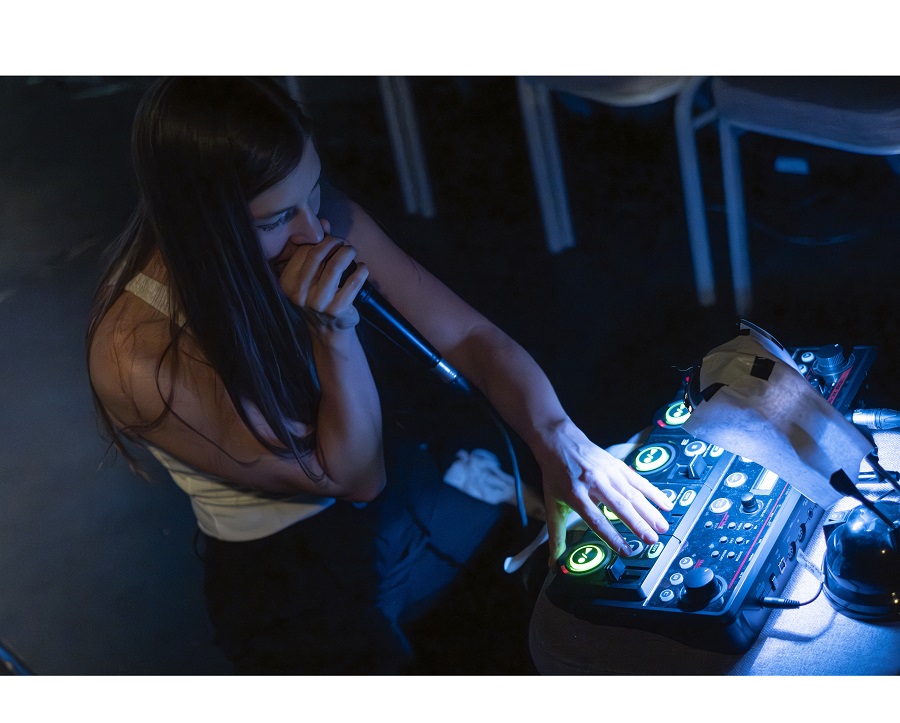 Na zdjęciu młoda dziewczyna siedzi przy konsoli didżejskiej, w ręce trzyma mikorofon, coś mowi.