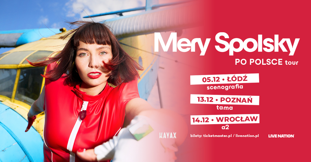 Kobieta zwrócona do obiektywu. Wyciąga dłoń. Ma krótkie brązowe włosy zakończone czerwonymi pasemkami. Ma na sobie czerwony kombinezon.