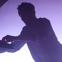 Mężczyzna na scenie pochyla się nad konsoletą didżejską. Jest ciemno, za nim widać tylko duży, fioletowy krąg światła.