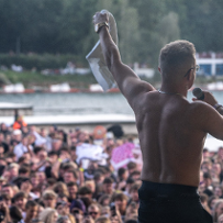 Mężczyzna stojący tyłem bez koszulki na scenie. Przed nim tłum ludzi.