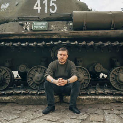 Mężczyzna w dżinsach i ciemnozielonym swetrze siedzi na gąsienicy zabytkowego czołgu zaplatając dłonie. Ma skupiony wyraz twarzy.