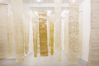 Na zdjęciu widoczna jest instalacja artystyczna składająca się z pionowo zawieszonych, półprzezroczystych tkanin w odcieniach bieli, beżu i żółci. Materiały te mają nieregularne struktury, które przypominają naturalne formy, takie jak wosk pszczeli czy pergamin. Instalacja ta buduje atmosferę kontemplacji i skupienia, symbolizując pracę pszczół i kolektywny charakter ich działań.