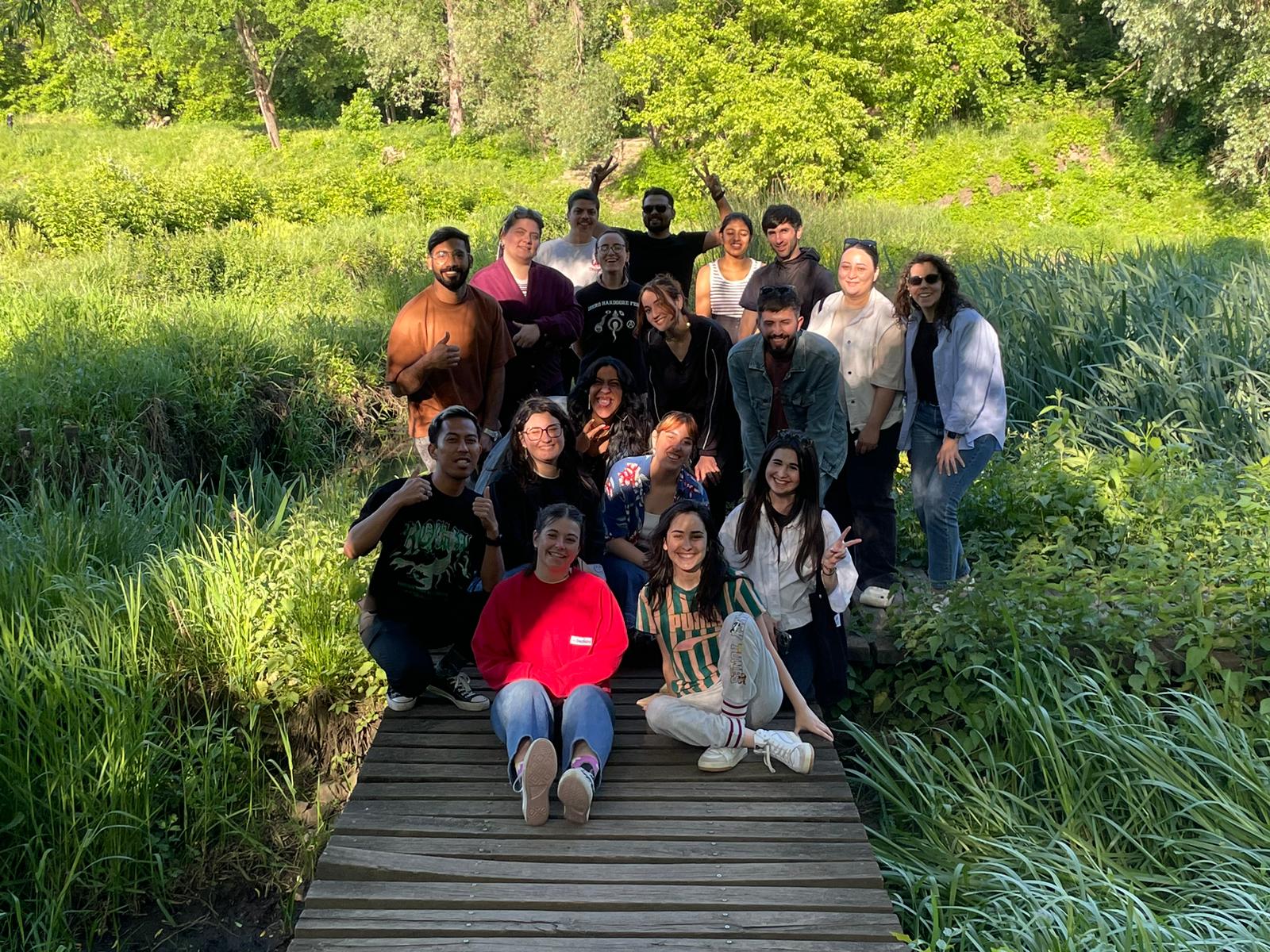 Grupa młodych ludzi stoi na drewnianym pomoście i pozuje do zdęcia. Wokół nich zielona łąka z wysokimi krzewami.