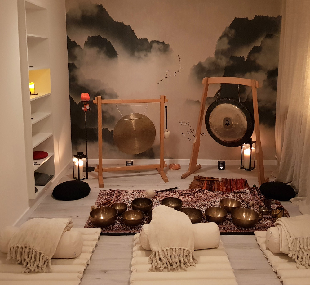 Zdjęcie kameralnej sali, w której stoją gongi i misy położone na dywanie. Na podłodze leżą białe karimaty, wałki i koce, dookoła stoją zapalone świece.