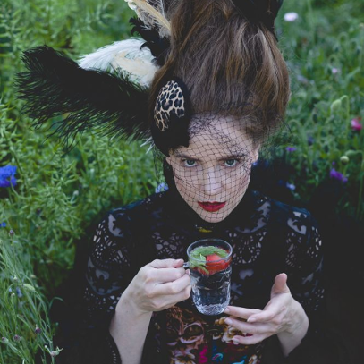 Młoda kobieta w czarnej, koronkowej sukni i toczku na głowie pije ze szklanki napar z truskawką i miętą.
