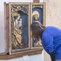 Młoda kobieta zwiedzająca wystawę pochyla się nad eksponatem - zamkniętym w gablocie drewnianym ołtarzykiem, na którym widnieje figura Matki Boskiej z Jezusem. Figura jest bogato zdobiona, pełna złoceń.