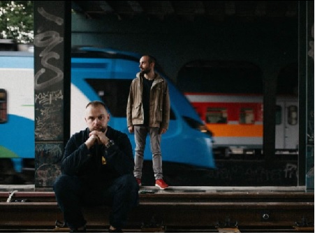 Mrokas stoi na peronie i Mrokas siedzi na peronie. W tle pociągi.