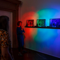 Na ścianie podświetlonej ostrymi światłami - czerwonym, zielonym, niebieskim, jest półka z trzema fotografiami, pozornie do siebie podobnymi. Patrzą na nie trzy zwiedzające.