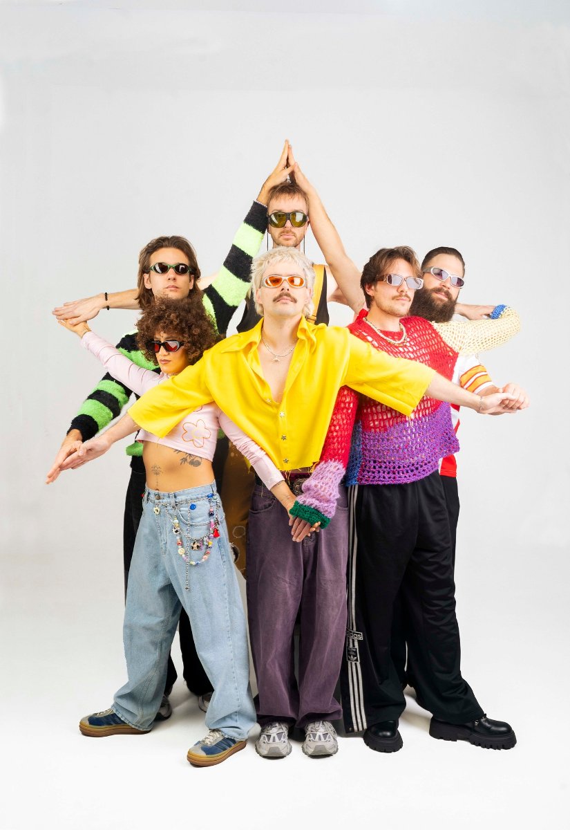 Sześcioosobowy zespół ubrany w kolorowe, eklektyczne stroje z lat 90. i okulary przeciwsłoneczne, ustawiony w symetrycznej pozie przypominającej totem lub rozłożone skrzydła na białym tle.