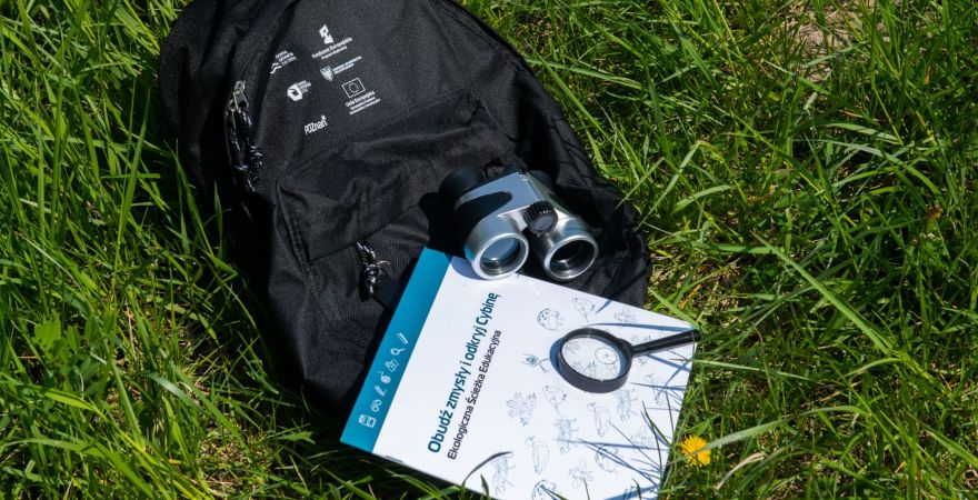 czarny pleacak leżący na trawie. Na nim lornetka, lupa i zeszyt.