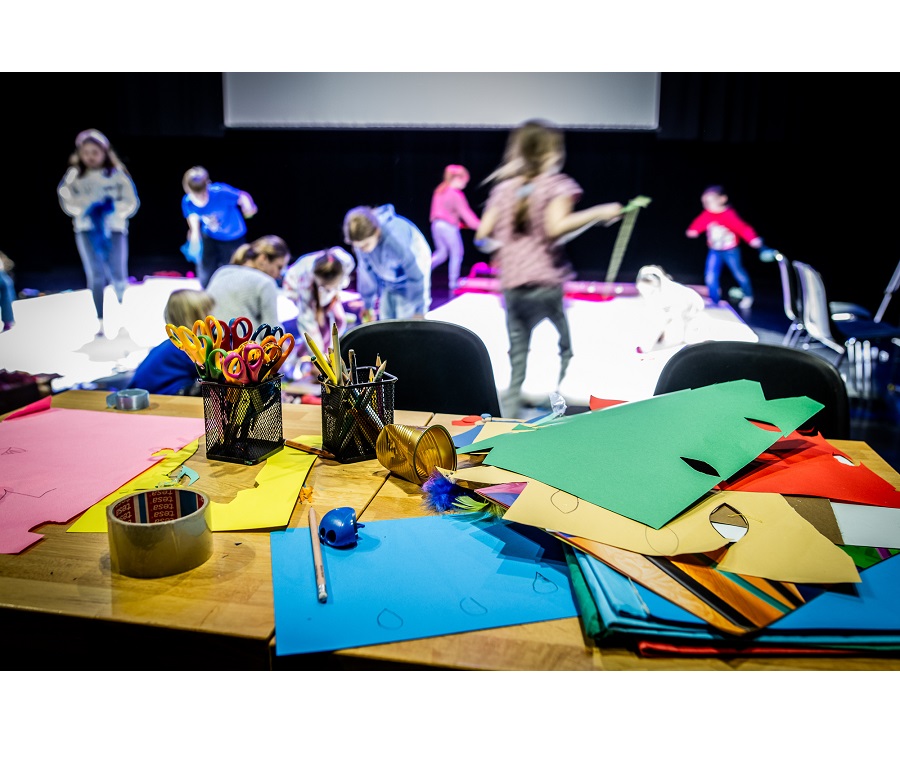 na zdjeciu grupa dzieci podczas zajęć. W tle widać siedzące na podłodze i biegajace dzieci. Na pierwszym planie stół z przyborami do zajęć plastycznych: kredkami, kolorowym papierem, nożyczkami.