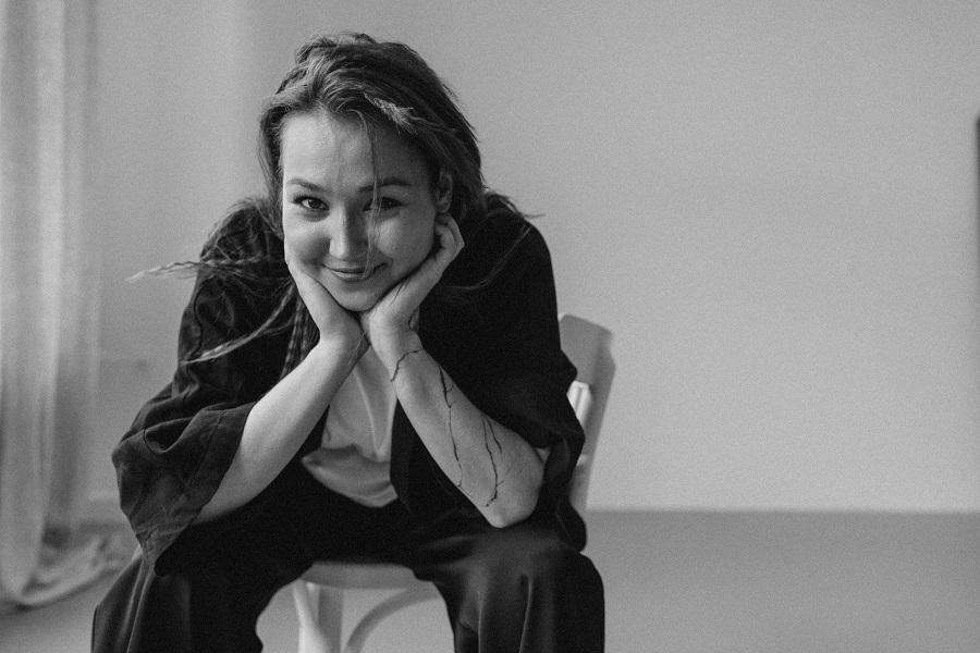 Zdjęcie młodej kobiety, która uśmiechnięta siedzi na krześle, rekoma podpiera głowę.