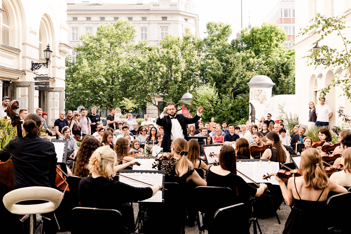 Zdjęcie orkiestry grającej na placu pomiędzy dwoma budynkami. Przed orkiestrą dyrygent, za dyrygentem publiczność, drzewa i budynek.